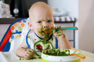 Baby in a highchair covered in green puree while learning to use a spoon